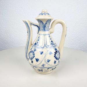 Op de afbeelding: Kleine, gebroken witte keramische kan met deksel, handvat en tuit. De kan is versierd met blauwe bloemen- en krulpatronen. Het handvat is gebogen en de tuit is slank. Het deksel heeft een kleine knop. De kan is waarschijnlijk voor het serveren van vloeistoffen.