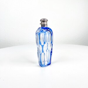 May include: A clear glass perfume bottle with a silver-colored cap and blue accents. The bottle has a geometric design with vertical blue lines and a rectangular shape. The bottle is displayed on a white surface.