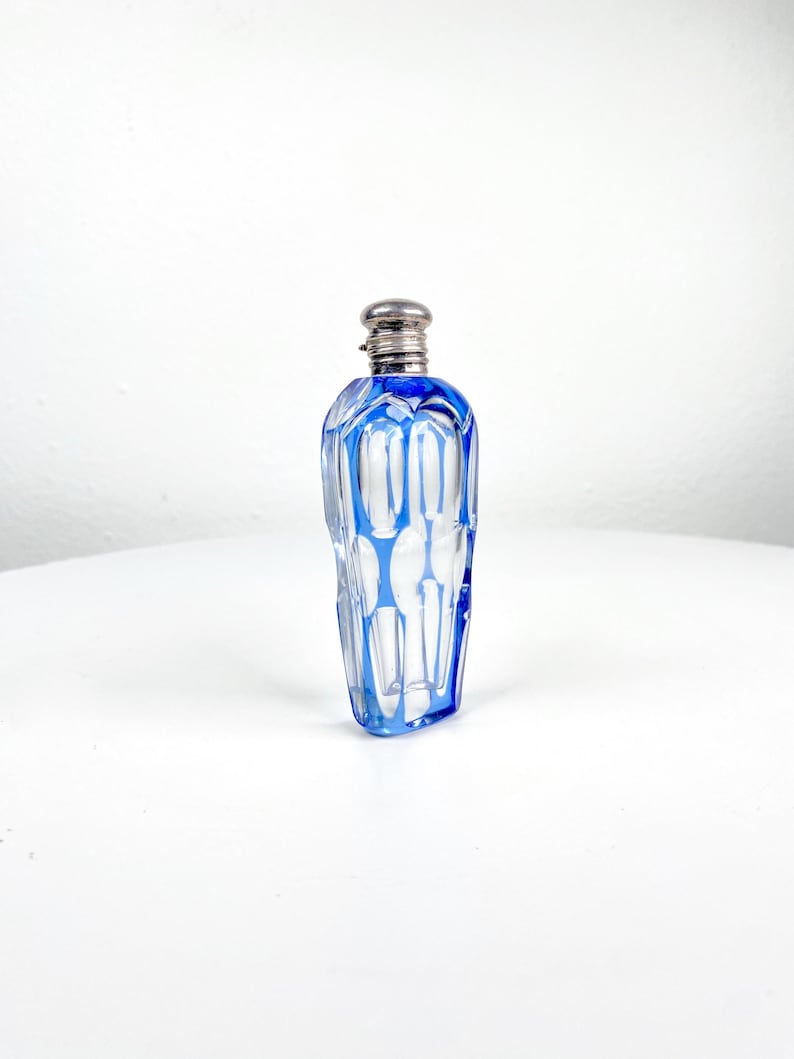May include: A clear glass perfume bottle with a silver-colored cap and blue accents. The bottle has a geometric design with vertical blue lines and a rectangular shape. The bottle is displayed on a white surface.