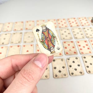 Op de afbeelding: Een vintage speelkaart met de Koning van Schoppen, vastgehouden in een hand. De kaart toont een gedetailleerde illustratie van een koning in rood, groen en goud. Andere speelkaarten zijn op de achtergrond, met rode harten en zwarte schoppen.