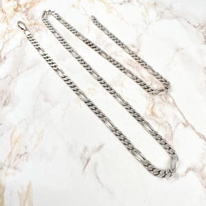 Op de afbeelding: Een zilverkleurige Figaro-ketting met een karabijnsluiting. De ketting ligt op een wit en grijs marmeren oppervlak. De ketting bestaat uit kleine, platte, ovale schakels die afwisselen met grotere, langwerpige schakels.