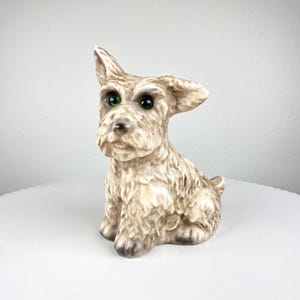 Könnte beinhalten: Keramikfigur eines sitzenden Hundes mit strukturiertem, hellbraunem Fell und grünen Augen. Der Hund hat ein aufgestelltes Ohr. Die Figur steht auf einer weißen Oberfläche.