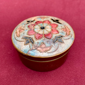 Op de afbeelding: Een rond koperen doosje met een deksel voorzien van cloisonné emaillewerk. Het deksel toont een centraal rood en wit bloemmotief omringd door lichtblauwe, zwarte en roze decoratieve elementen op een getextureerde, lichtblauwe achtergrond.