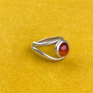 Op de afbeelding: Een zilveren ring met een ronde, rood-oranje steen. De ring heeft een uniek ontwerp met een gesplitste band die om de steen buigt. De ring staat tegen een gele achtergrond.
