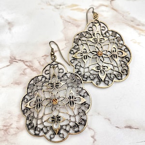 Op de afbeelding: Een paar gouden filigraan oorbellen met een kleine, heldere strasssteen in het midden van elke oorbel.