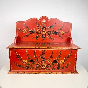 Op de afbeelding: Een rechthoekige houten wanddoos met een scharnierend deksel, geschilderd in een levendige rode kleur. De voor- en achterpanelen zijn voorzien van handgeschilderde bloemmotieven in zwart, geel en groen, met een kleine vogel op het voorpaneel.