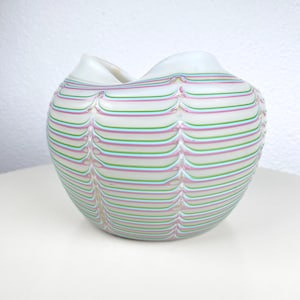 Op de afbeelding: Een witte glazen vaas met een brede, golvende rand. De vaas is versierd met horizontale strepen in roze, groen en lichtblauw. De strepen zijn gelijkmatig verdeeld en creëren een textuureffect. De vaas staat op een wit oppervlak.