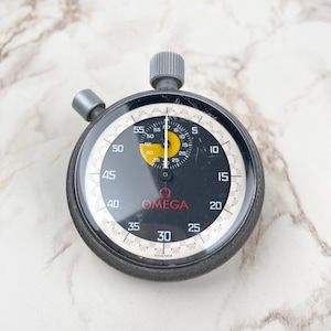 Op de afbeelding: Een zwarte en zilveren Omega stopwatch met een witte en zwarte wijzerplaat. De wijzerplaat heeft witte cijfers en markeringen, met een gele kern en het Omega-logo in rood. De stopwatch heeft een grijze opwindknop en een zijknop.