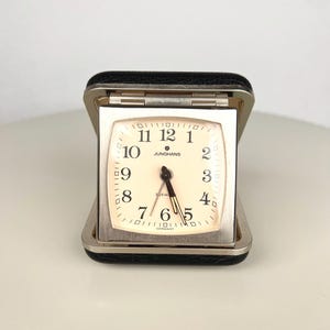 May include: A vintage Junghans travel alarm clock in a black textured folding case. The square clock face features a cream background, black Arabic numerals, dark hands, and the text Junghans, 2 Jewels, and Germany printed on the dial.