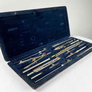 Puede incluir: Una caja de cuero vintage con un interior de terciopelo azul contiene un juego de herramientas de dibujo, que incluyen compases, reglas y lápices.