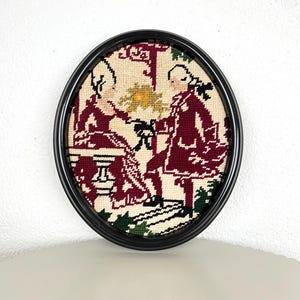 Puede incluir: Arte de tapiz enmarcado ovalado que representa dos figuras con atuendo burdeos, sobre un fondo crema. La obra de arte presenta un marco negro y una escena detallada con acentos verdes y amarillos, que muestra un tema histórico.
