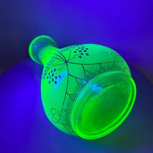 May include: A vibrant green glass vase, illuminated by a black light, showcasing a neon effect. The vase features a rounded body and a flared rim, with a detailed design of leaves and berries. The vase is a decorative item.