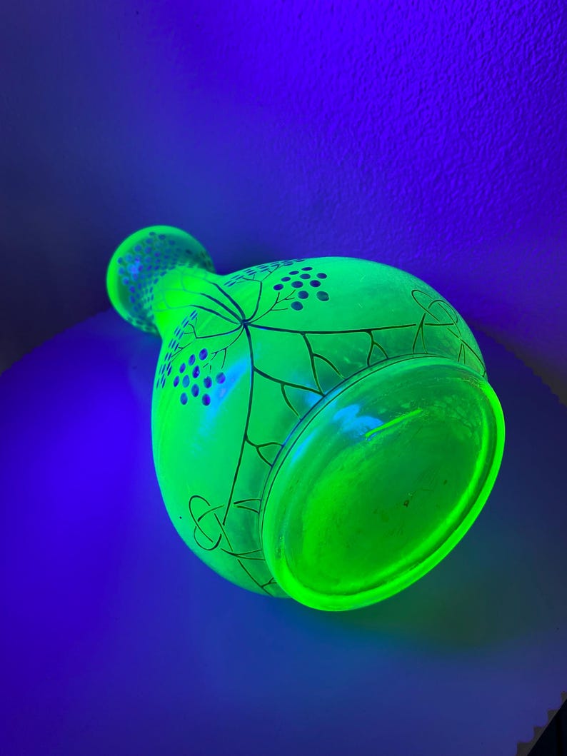 May include: A vibrant green glass vase, illuminated by a black light, showcasing a neon effect. The vase features a rounded body and a flared rim, with a detailed design of leaves and berries. The vase is a decorative item.