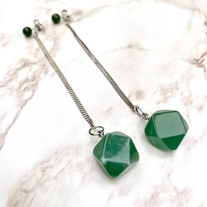 Op de afbeelding: Een paar lange zilveren kettingoorbellen met groene edelsteen hangers. De edelstenen zijn gefacetteerd en hebben een geometrische vorm.