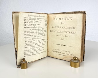 Op de afbeelding: Een open antiek boek getiteld "ALMANAK der VADERLANDSCHE GESCHIEDENISSEN voor het Jaar 1816" met tekst in het Nederlands. Twee kleine messing inktpotten staan op het witte oppervlak. De pagina's van het boek zijn vergeeld en de kaft is versleten.