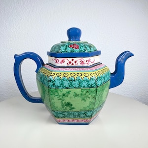 Könnte beinhalten: Eine farbenfrohe Keramik-Teekanne mit blauem Griff, Ausguss und Deckel. Der Deckel hat ein rotes Blumenmuster. Der Körper der Teekanne ist mit grünen, gelben und rosa Mustern verziert. Die Teekanne steht auf einer weißen Oberfläche.