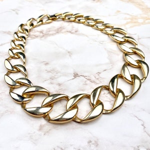 Op de afbeelding: Een goudkleurige ketting met grote, gepolijste, in elkaar grijpende ovale schakels. De ketting is in een cirkelvorm gerangschikt en toont een gladde, reflecterende metalen afwerking op elke individuele schakel die de doorlopende ketting vormt.