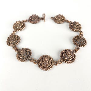 Puede incluir: Un brazalete vintage de tono cobrizo con eslabones de filigrana circulares y ornamentados. Cada eslabón presenta un diseño detallado de puntos en relieve y patrones arremolinados, conectados por pequeñas cadenas. El brazalete tiene un cierre de gancho.
