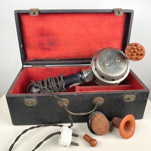 May include: An antique black case with a red velvet interior holds a vintage massage device. The device is silver and black with a brown textured head. Additional brown and black massage attachments are displayed outside the case.