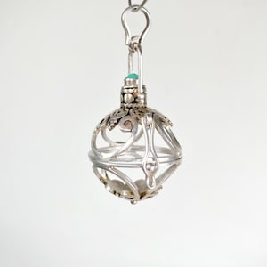 May include: A silver filigree pendant with a small turquoise stone. The pendant is spherical with an openwork design and a decorative top. It hangs from a silver hook.