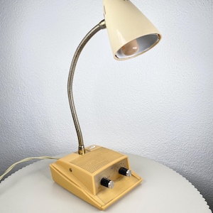 May include: A vintage desk lamp with a cream-colored conical shade and a flexible, silver-toned gooseneck. The base is a light yellow-beige with two black knobs and a power switch. The lamp is plugged into a white cord.