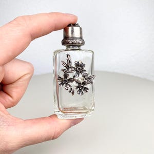 Puede incluir: Pequeño frasco de perfume de vidrio transparente con un diseño floral plateado. El frasco tiene una tapa plateada y está sostenido por una mano. El diseño floral presenta flores y hojas detalladas.