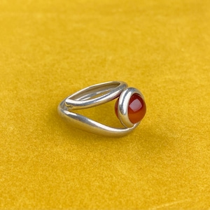 Op de afbeelding: Een zilveren ring met een rode carneol-edelsteen. De ring heeft een uniek ontwerp met een gebogen band die de steen omsluit.