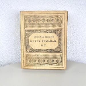 Op de afbeelding: Een vintage boek met een bruinachtige kaft met de tekst "NEDERLANDSCHE MUZEN-ALMANAK, 1826." De kaft heeft decoratieve randen en patronen in een donkerdere bruintint. Het boek is rechthoekig en lijkt in goede staat te verkeren.