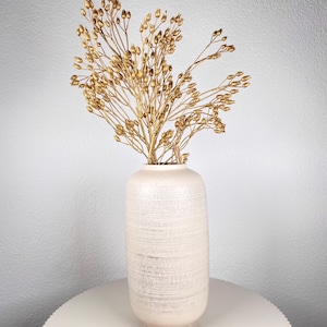 May include: A tall, off-white ceramic vase with a textured, horizontal stripe design. The vase holds a bouquet of dried, golden-colored ornamental grass. The vase is displayed on a round, white table against a white wall.