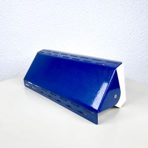 Op de afbeelding: Een moderne, blauwe metalen wandlamp met een rechthoekige vorm. De lamp heeft een glanzende afwerking en een reeks horizontale sleuven langs de bovenrand. De achterkant van de lamp is wit.