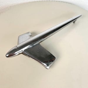May include: A polished chrome automotive hood ornament shaped like a stylized jet or rocket. The object features a pointed nose, two swept-back wings, and a long, tapered tail section. The metallic surface reflects light, showing a smooth finish.