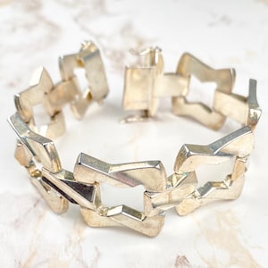 Op de afbeelding: Een zilveren armband met een geometrisch ontwerp. De armband is gemaakt van in elkaar grijpende schakels die de vorm hebben van vierkanten met uitsparingen.