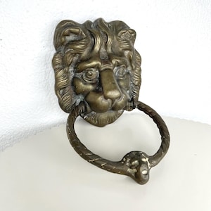 Könnte beinhalten: Antiker Türklopfer mit Löwenkopf aus Messing mit Ringgriff. Das Gesicht des Löwen weist detaillierte Merkmale auf, darunter Mähne, Nase und Augen. Der Ringgriff ist verdreht und am Maul des Löwen befestigt.