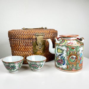 Op de afbeelding: Een verzameling antieke, op Azië geïnspireerde items, waaronder een geweven mand met een messing sluiting, een decoratieve theepot en twee kleine theekopjes. De theepot en theekopjes hebben kleurrijke bloemen- en figuurontwerpen.