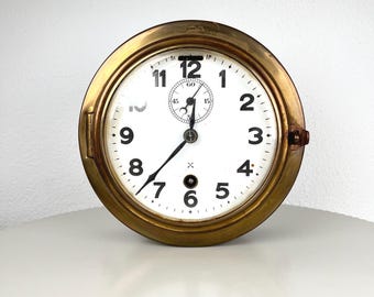 Op de afbeelding: Een ronde, antieke klok met een messingkleurig frame en een witte wijzerplaat. Zwarte cijfers markeren de uren, met een kleinere wijzerplaat bovenaan. De klok heeft zwarte wijzers en een kleine, bruine knop aan de zijkant.