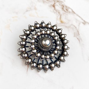 Puede incluir: Un broche de plata con un diseño circular que presenta un elemento central redondo rodeado de círculos concéntricos de elementos redondos más pequeños.