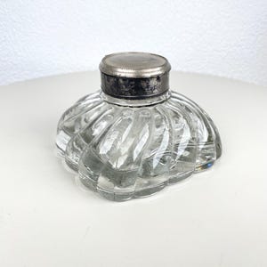 Puede incluir: Tintero de cristal transparente con tapa de metal plateado. El tintero tiene un diseño acanalado y festoneado. La tapa circular se asienta sobre el tintero. El tintero está sobre una superficie blanca.