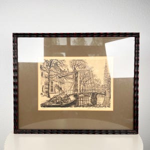 May include: A framed black and white ink drawing depicting a canal scene with a drawbridge, trees, and buildings. The artwork is set behind glass with a wide brown matting and enclosed in a dark, textured frame with subtle red highlights.