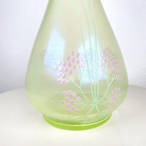 May include: A pale green glass vase with an iridescent finish. The vase is decorated with pink floral clusters and green stems. It has a rounded body and a flared base. The background is white.