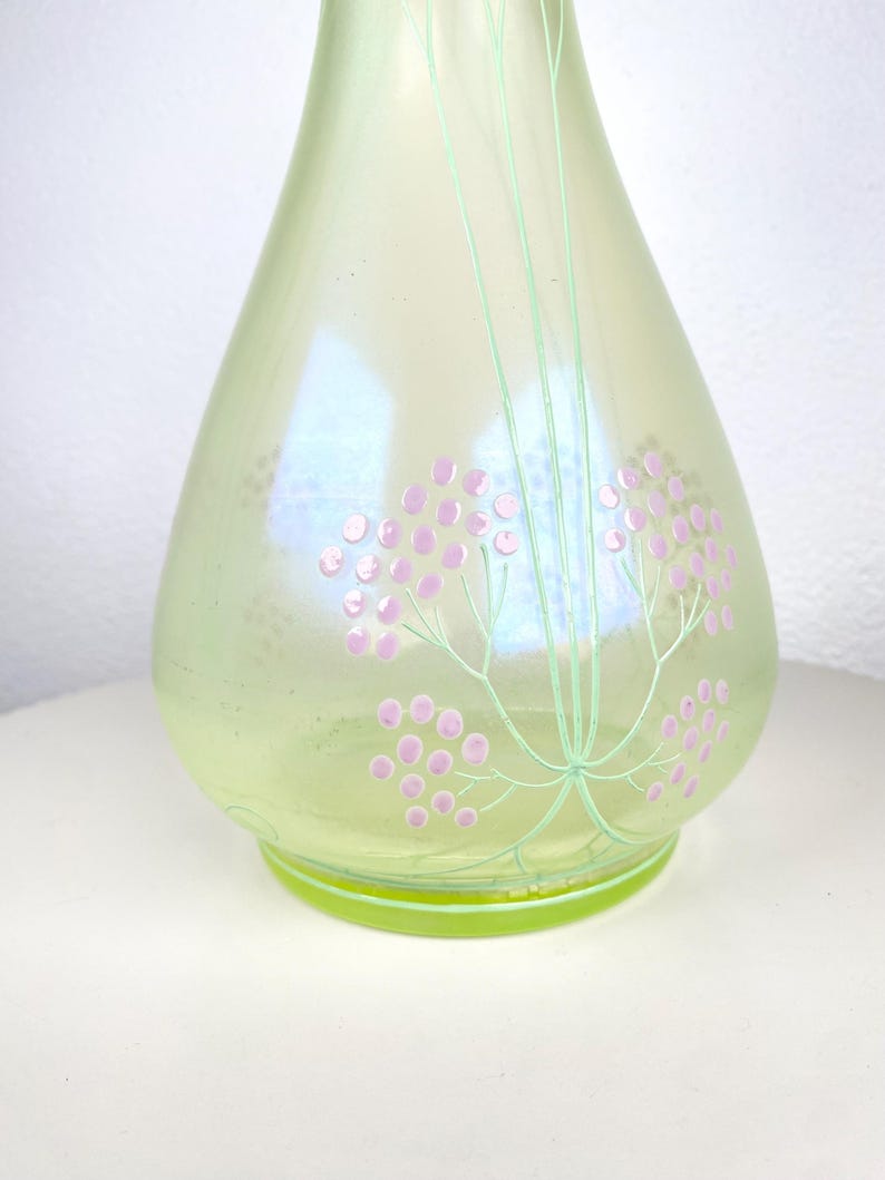 May include: A pale green glass vase with an iridescent finish. The vase is decorated with pink floral clusters and green stems. It has a rounded body and a flared base. The background is white.