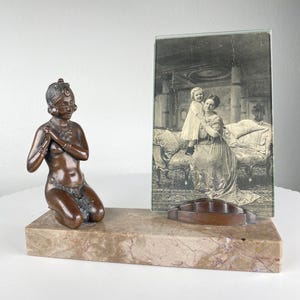 Puede incluir: Una escultura en bronce de una figura arrodillada con las manos juntas, junto a una fotografía en blanco y negro enmarcada. La escultura y la foto están sobre una base rectangular de mármol con un objeto geométrico marrón.