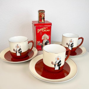 Peut inclure: Un ensemble de trois tasses et soucoupes en céramique aux tons crème et rouge foncé, orné d'une infirmière tenant une boîte de cacao Droste's. Derrière les tasses se trouve une boîte métallique rectangulaire rouge de cacao Droste's assortie.