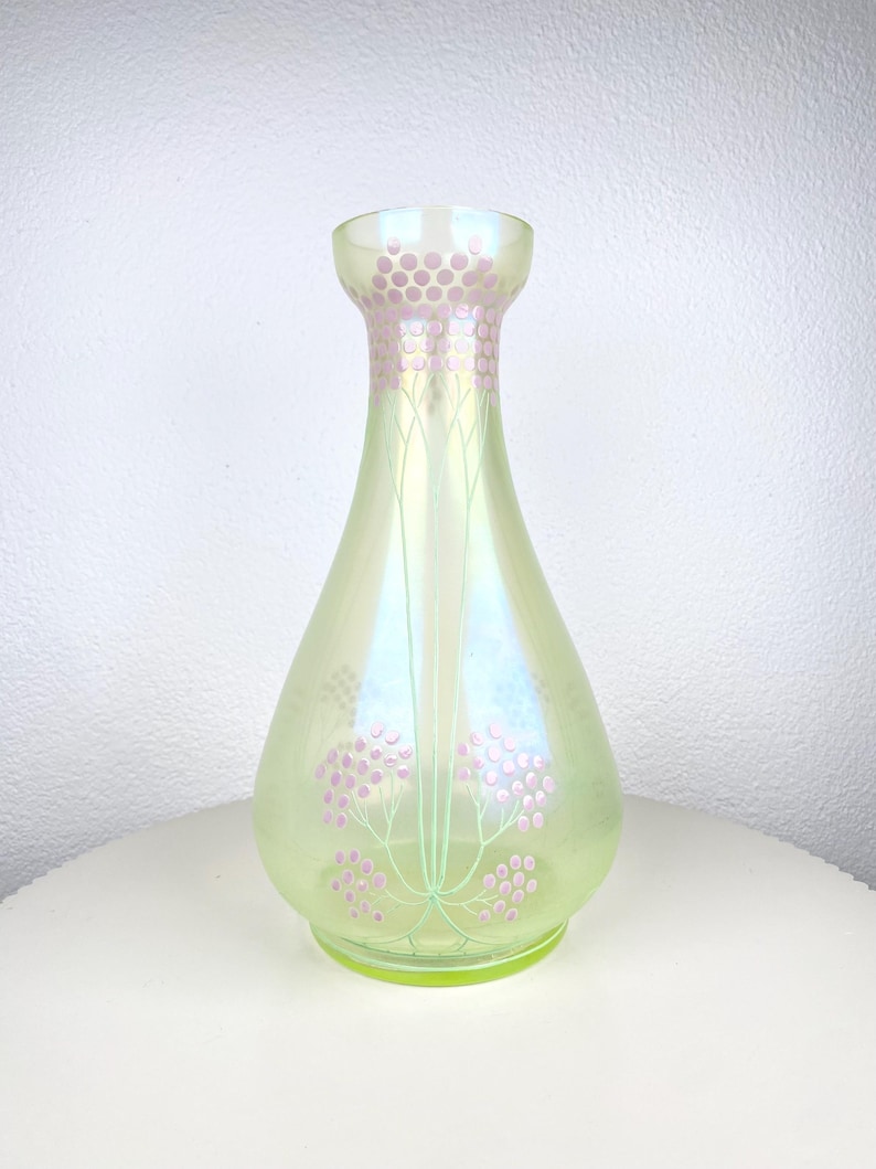 May include: An iridescent glass vase in a pear shape, with a light green colour. It has a decorative pattern of pink floral designs and a matching pink dotted pattern around the neck. The vase is on a white surface.