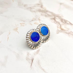 Op de afbeelding: Een paar ronde oorknopjes met een centrale ronde blauwe steen met subtiele gouden spikkels, gezet in een gepolijst zilveren frame met een decoratief, stralend geribbeld of geschulpt randontwerp rondom de omtrek.