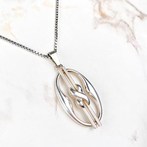 Op de afbeelding: Een zilveren ketting met een hanger met een gestileerd knoopontwerp in een ovaal frame.