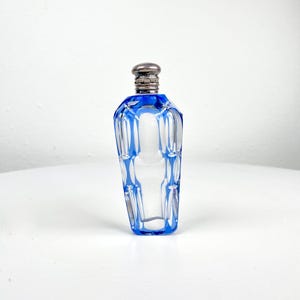 May include: A clear glass perfume bottle with a blue geometric design and a silver-colored cap. The bottle has a faceted shape and is displayed on a white surface. The bottle is approximately 4 inches tall.