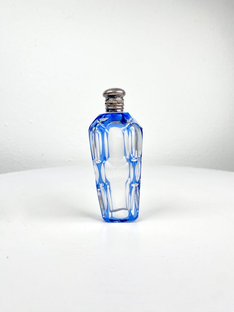 May include: A clear glass perfume bottle with a blue geometric design and a silver-colored cap. The bottle has a faceted shape and is displayed on a white surface. The bottle is approximately 4 inches tall.