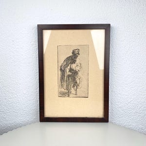 Op de afbeelding: Een ingelijste zwart-wit ets van een oudere persoon die op een wandelstok leunt. Het kunstwerk is geplaatst tegen een crèmekleurige achtergrond in een donkerbruine houten lijst. De afbeelding wordt weergegeven op een wit oppervlak.