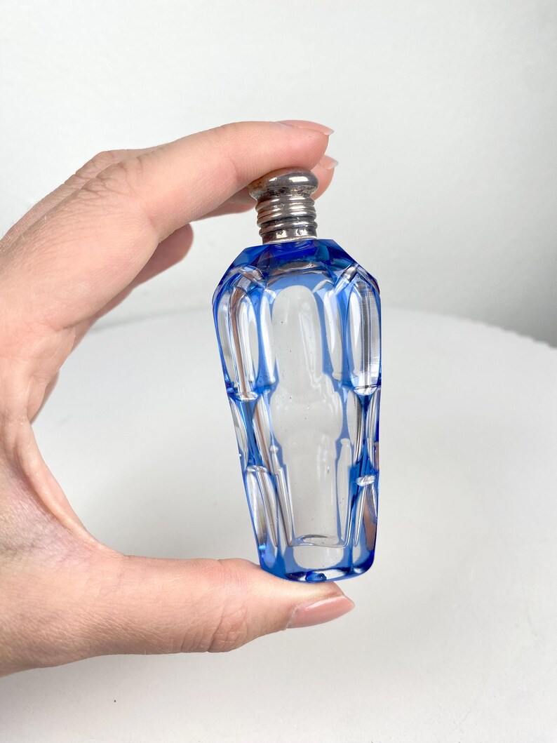 May include: A small, clear glass perfume bottle with a blue geometric design. The bottle has a silver-colored metal cap. The bottle is held in a hand against a white background.