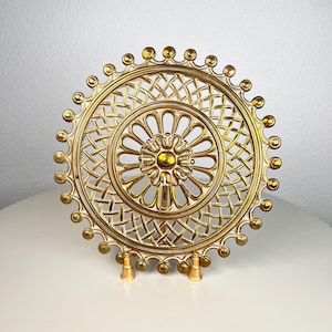 May include: A circular gold-toned metal decorative plate featuring an intricate lattice pattern, a central floral motif, and a scalloped edge adorned with small raised spheres. The plate is supported by two small, matching gold-toned metal feet.